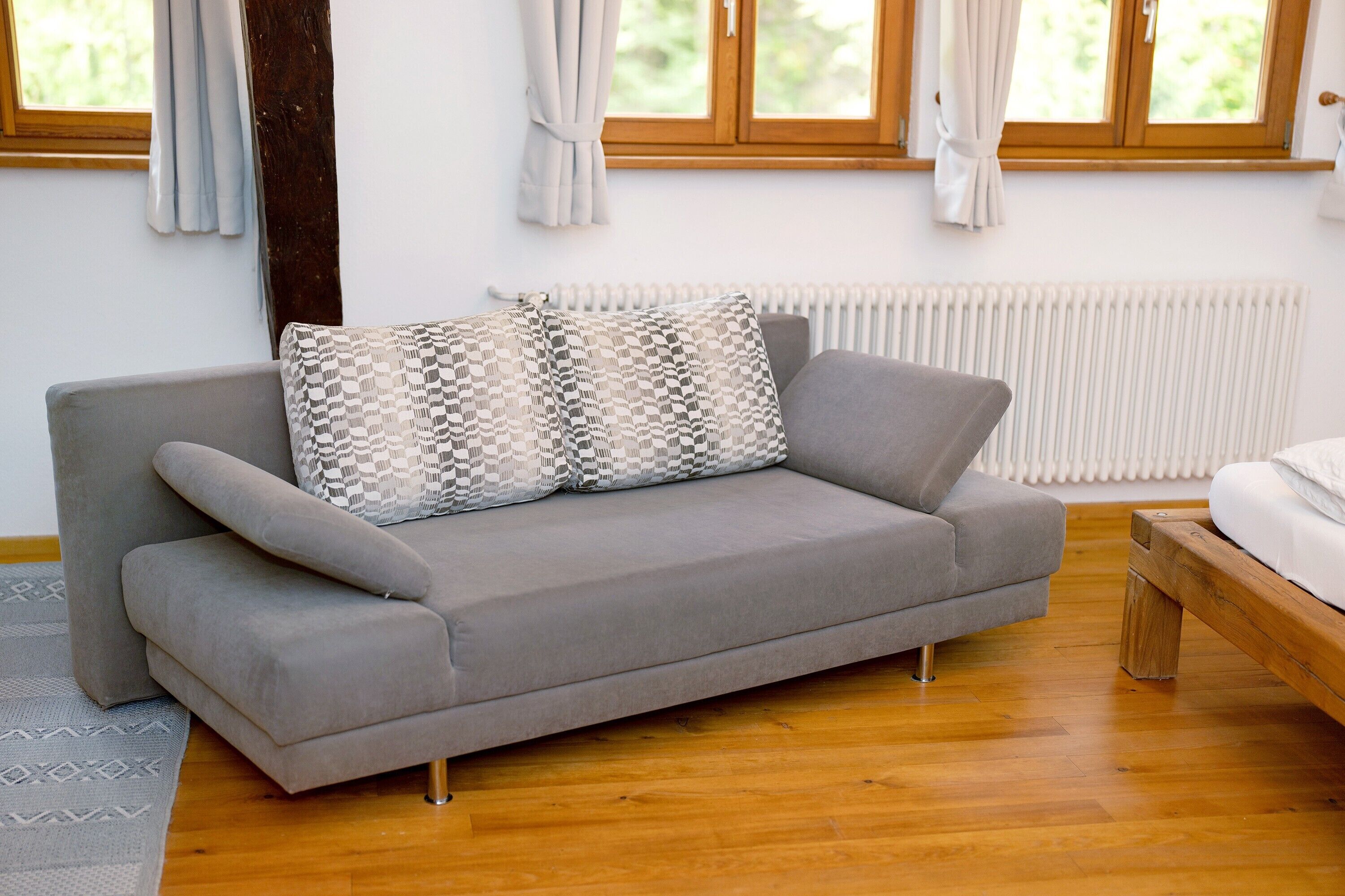 A grey sofa with patterned cushions, a wooden floor, and a window with curtains.