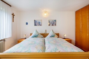 Un dormitorio con una cama de madera, dos mesitas de noche con lámparas, una cruz en la pared y dos cuadros enmarcados.