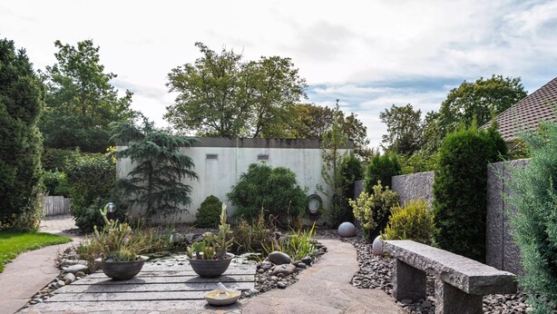 Un jardín con un sendero empedrado, plantas en macetas, un banco de piedra y una pared blanca con una ventana.
