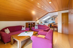 Un salon avec un plafond en bois, deux canapés rouges, une table basse en bois et, au fond, une cuisine.
