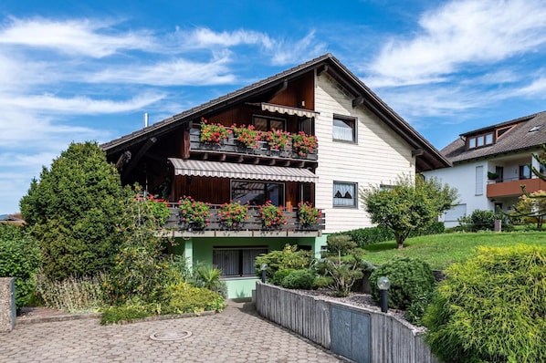 Une maison à deux étages avec un balcon, entourée de verdure et dotée d’une allée pavée.