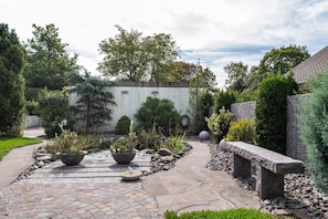 Un jardin avec un chemin pavé, des plantes en pot, un banc en pierre et un mur blanc agrémenté d’une fenêtre.