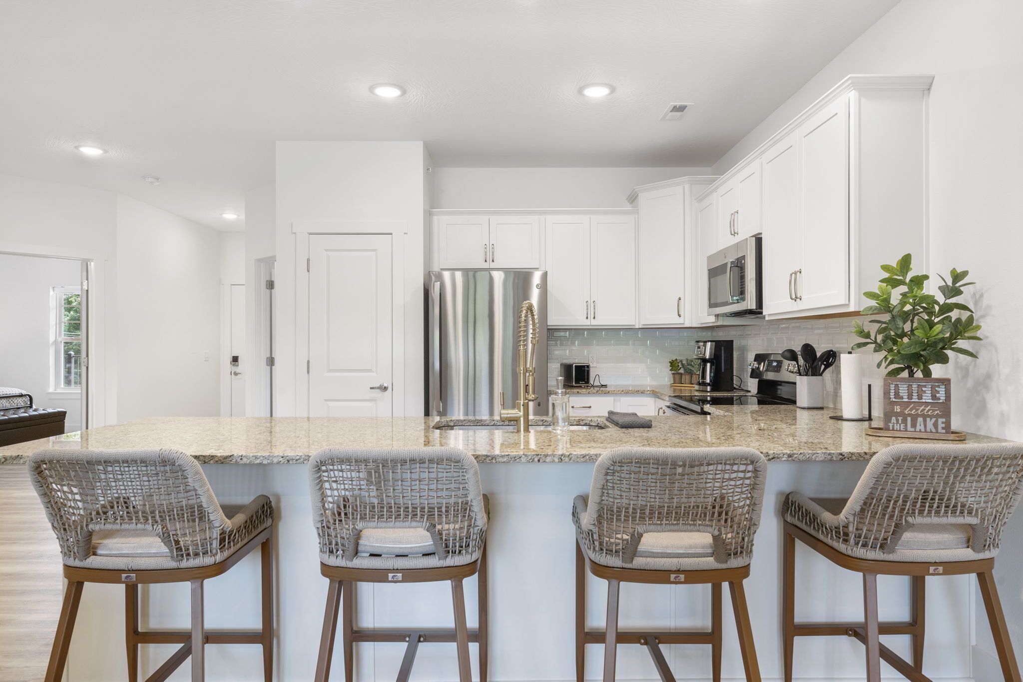 A modern kitchen with white cabinets, a stainless steel refrigerator, a microwave, and a coffee maker. There are four wicker bar stools and a potted plant.