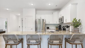 A modern kitchen with white cabinets, a stainless steel refrigerator, a microwave, and a coffee maker. There are four wicker bar stools and a potted plant. - Lakefront Retreat on Lake Taneycomo, Near Branson Landing and Attractions (Branson)
