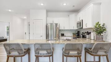 A modern kitchen with white cabinets, a stainless steel refrigerator, a microwave, and a coffee maker. There are four wicker bar stools and a potted plant.