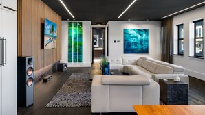 Ein modernes Wohnzimmer mit einem weißen Sofa, einem Couchtisch und einer Wand im Holzoptik. An den Wänden hängen zwei große Gemälde und auf der rechten Seite ein großes Fenster.