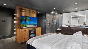 Ein modernes Hotelzimmer mit einem großen Bett, einem an einer Holzplatte montierten Fernseher, einer Glasdusche und einem Badezimmer mit einem großen Spiegel.
