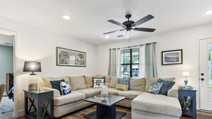 A living room with a sofa, two side tables, a coffee table, a ceiling fan, and a window with curtains.