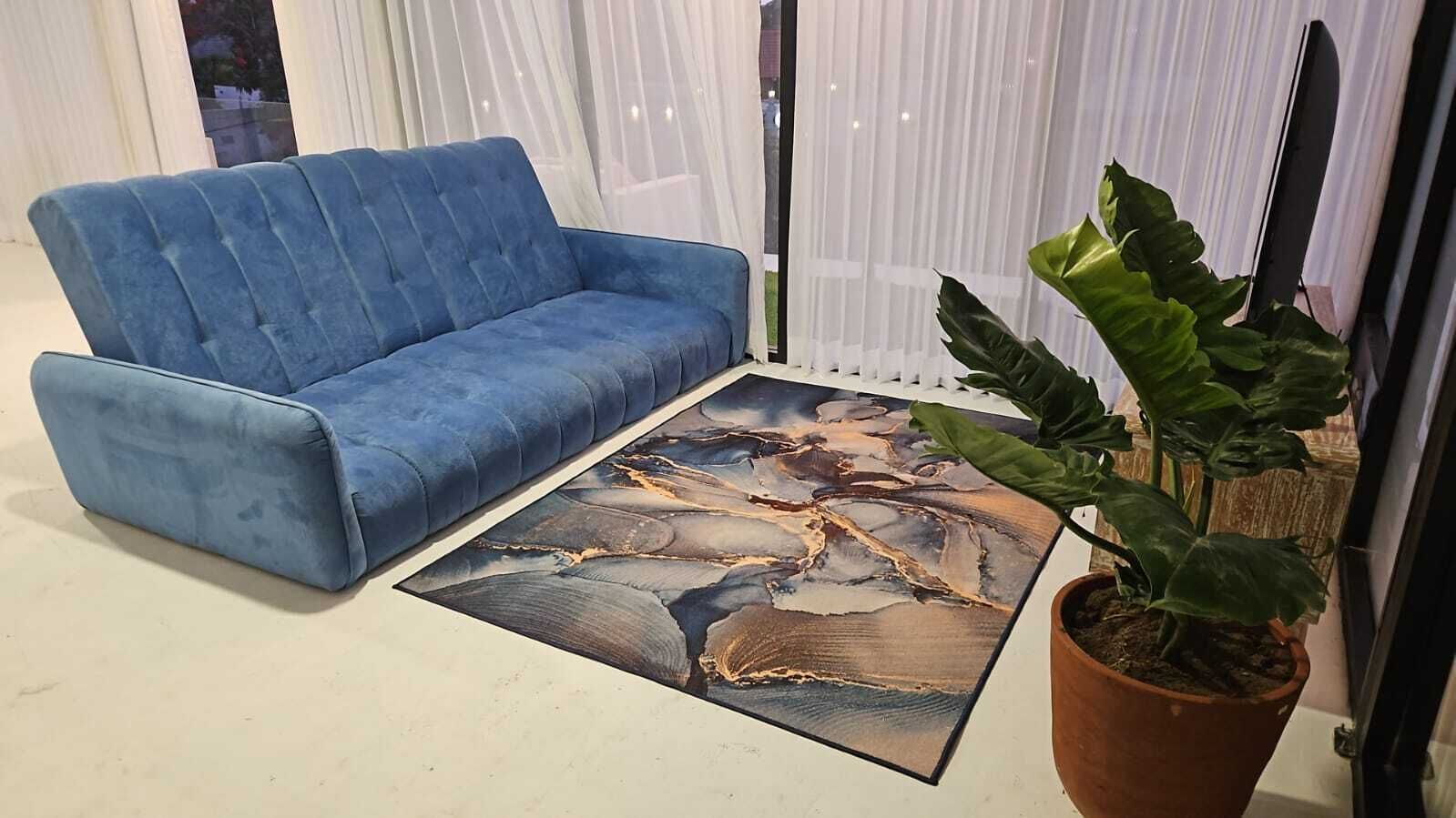 Sininen sohva, kuviollinen matto ja ruukussa oleva kasvi huoneessa, jossa on läpikuultavat verhot.
