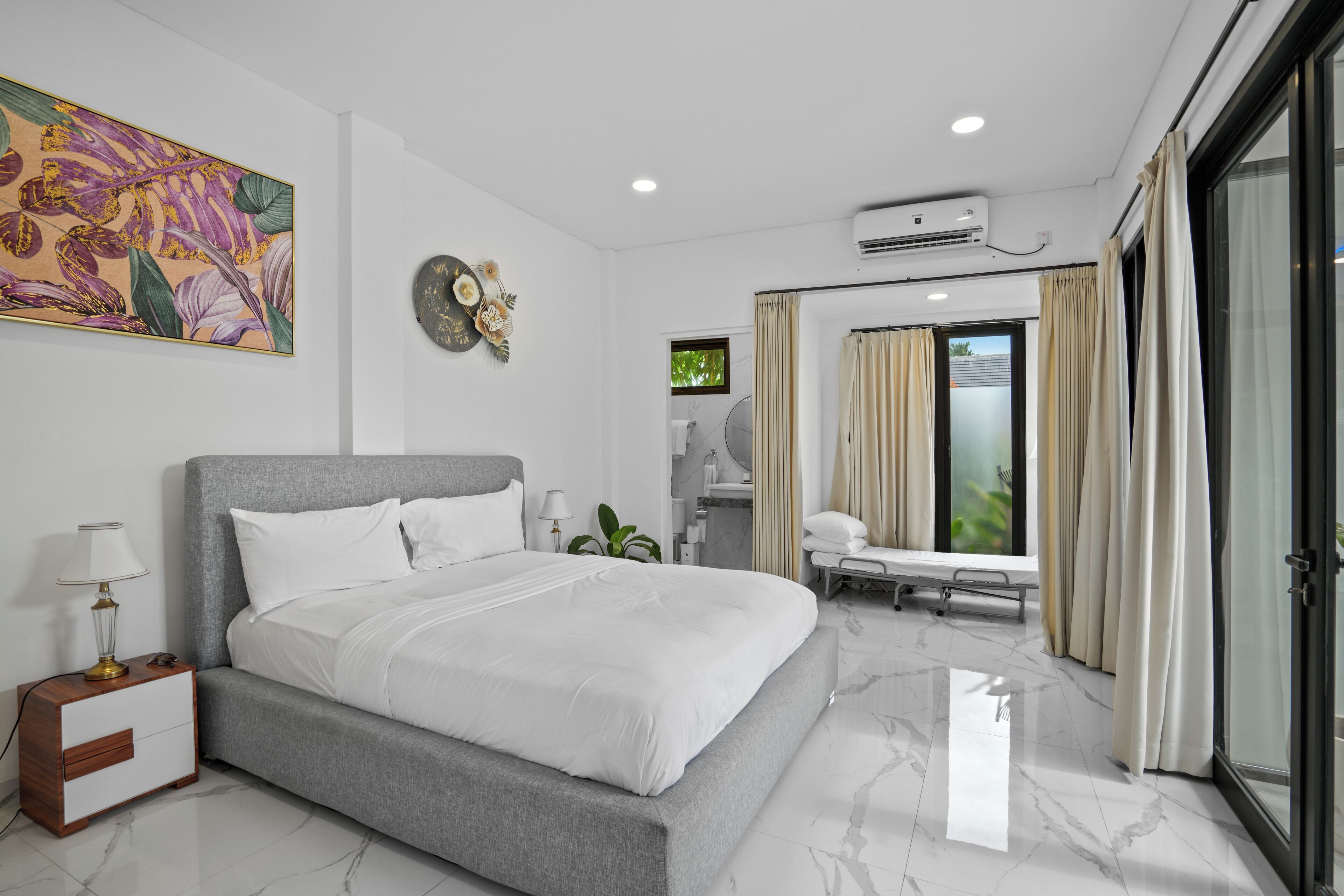 A modern hotel room with a large bed, a bedside table, a desk, and a chair. There is a window with curtains, an air conditioning unit, and a wall-mounted artwork.