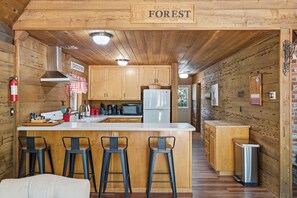 A wooden interior with a kitchen area featuring a central island with bar stools, wooden cabinets, and a refrigerator. - Shaver Wood Forrest-Smokey Tree (Shaver Lake)