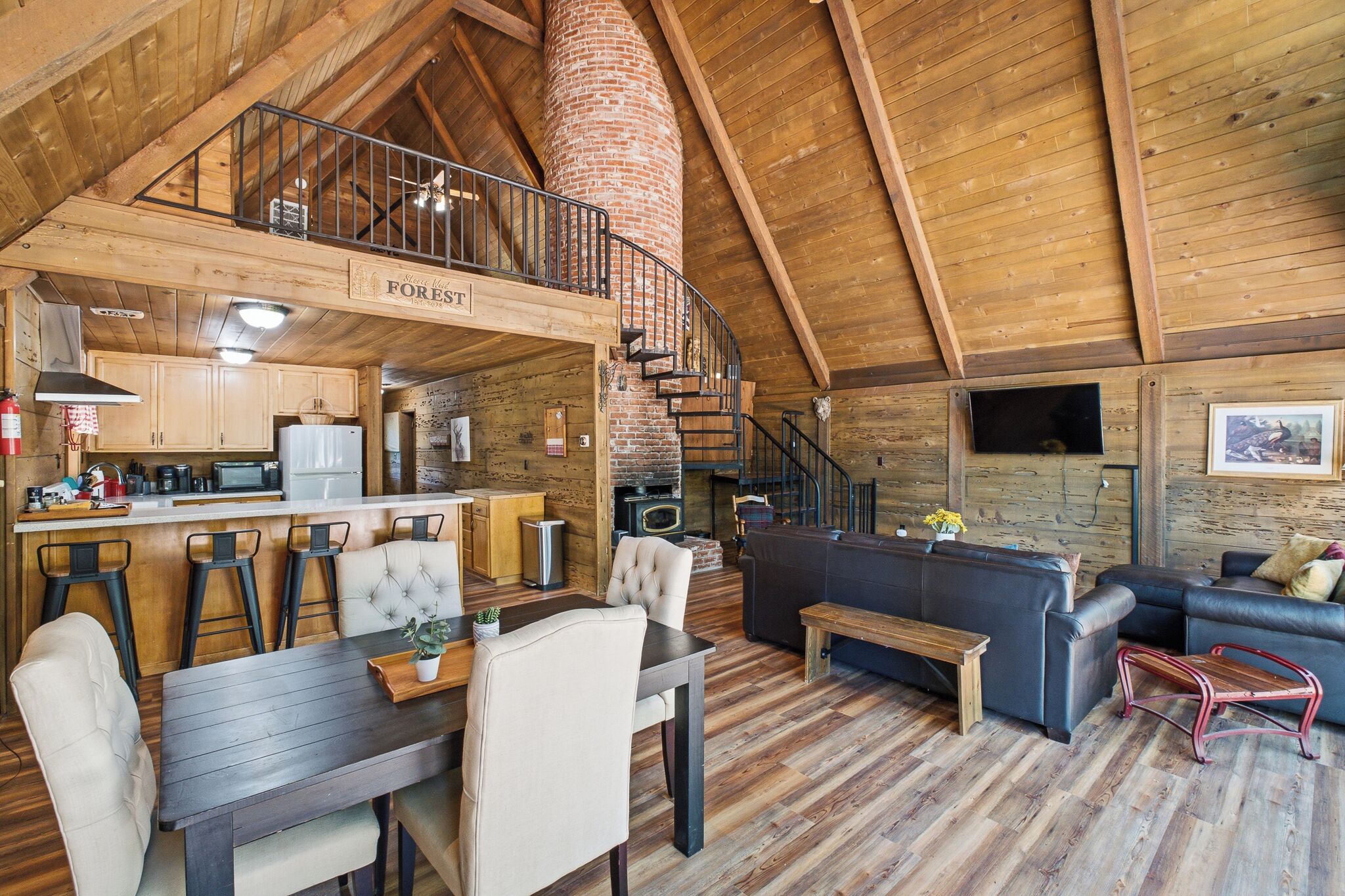 Een rustiek houten interieur met een eethoek, keuken en woonruimte, met zichtbare houten balken, een bakstenen schoorsteen en een slaaploft.