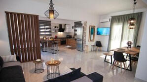 A modern living room with a sofa, a dining area with a table and chairs, a kitchen with a refrigerator and a microwave, and a TV mounted on the wall.