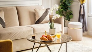 Ein gemütliches Wohnzimmer mit einem beigefarbenen Sofa, einem runden Couchtisch mit Croissants, einem Glas Orangensaft und einem kleinen Blumentopf.