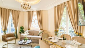 A well-lit room with a sofa, dining table set for four, and a chandelier. - Merveil - Luxury Suite - Louvre - Ours I (Paris)