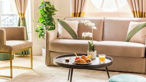 A cozy living room with a beige sofa, a round coffee table with pastries and a drink, a green plant, and a chair. - Merveil - Luxury Suite - Louvre - Ours I (Paris)