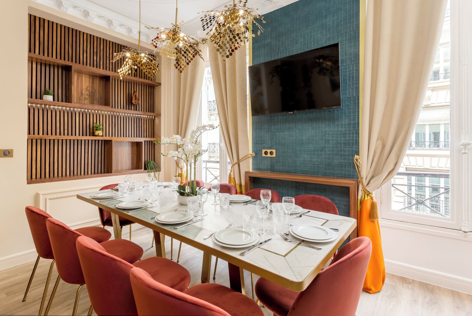 A dining area with a long table set for six, surrounded by red chairs, with a wooden panel wall and a TV mounted above.