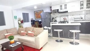 A modern living room with a sofa, a coffee table, and a small plant. There is a kitchen area with a refrigerator, microwave, and bar stools.