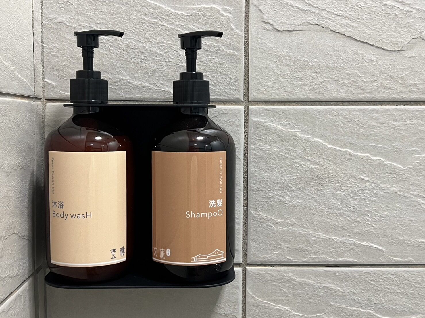Two bottles of body wash and shampoo on a black shelf against a textured wall.