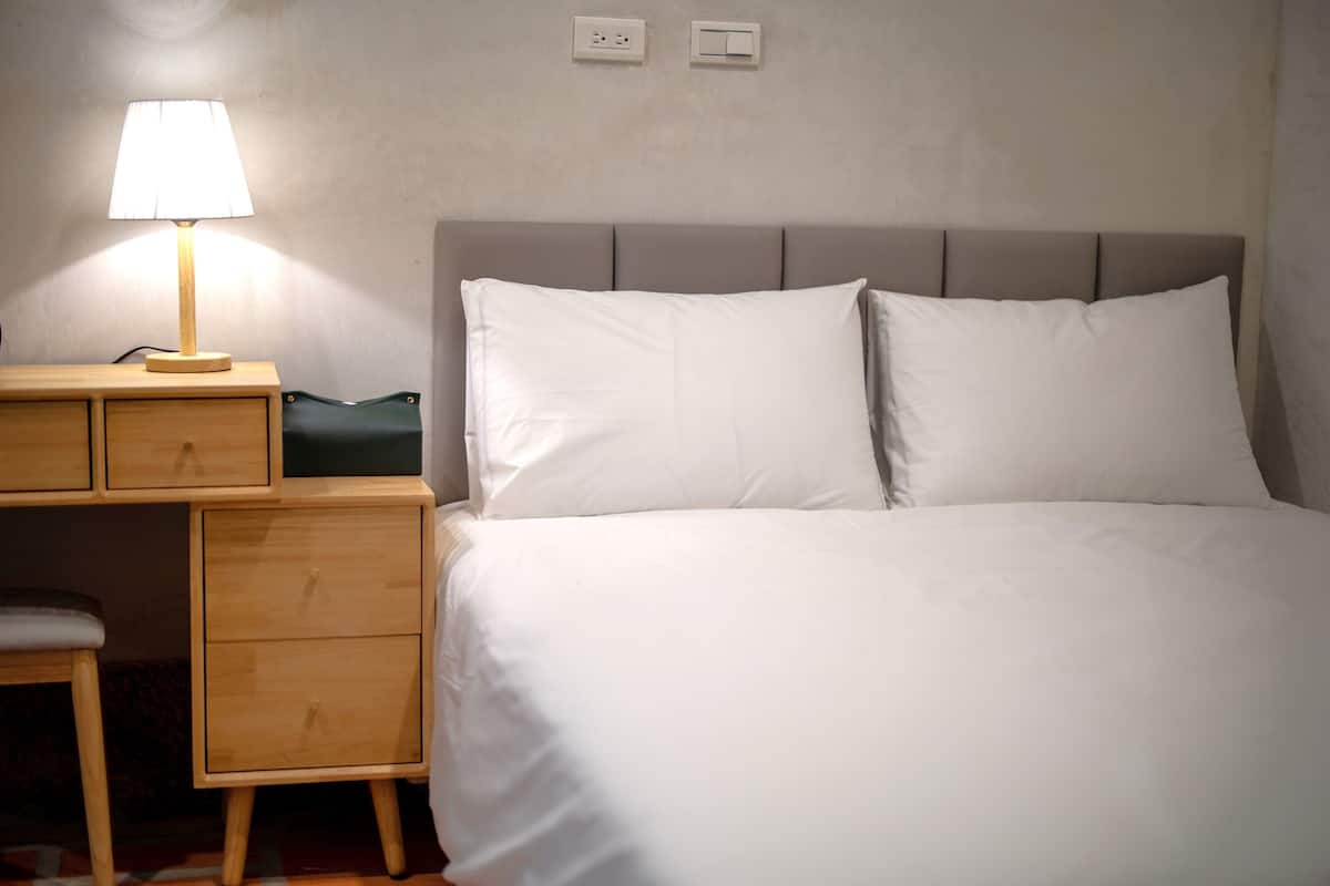 A neatly made bed with white linens, a bedside table with a lamp, and a small wooden nightstand with drawers.