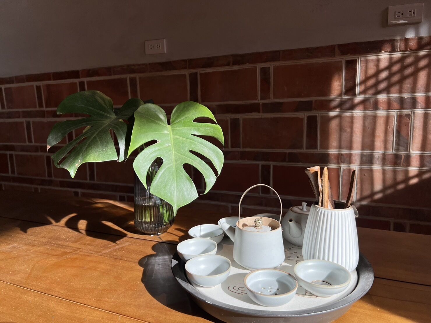 A table with a white tray holding several white bowls, a white teapot, and a white vase with a wooden handle. There is a large green plant in a glass vase on the table.