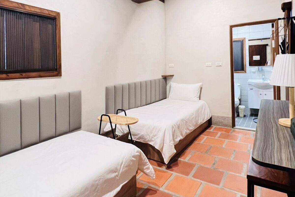 A small, cozy room with two beds, a small table, and a bathroom visible through an open door.