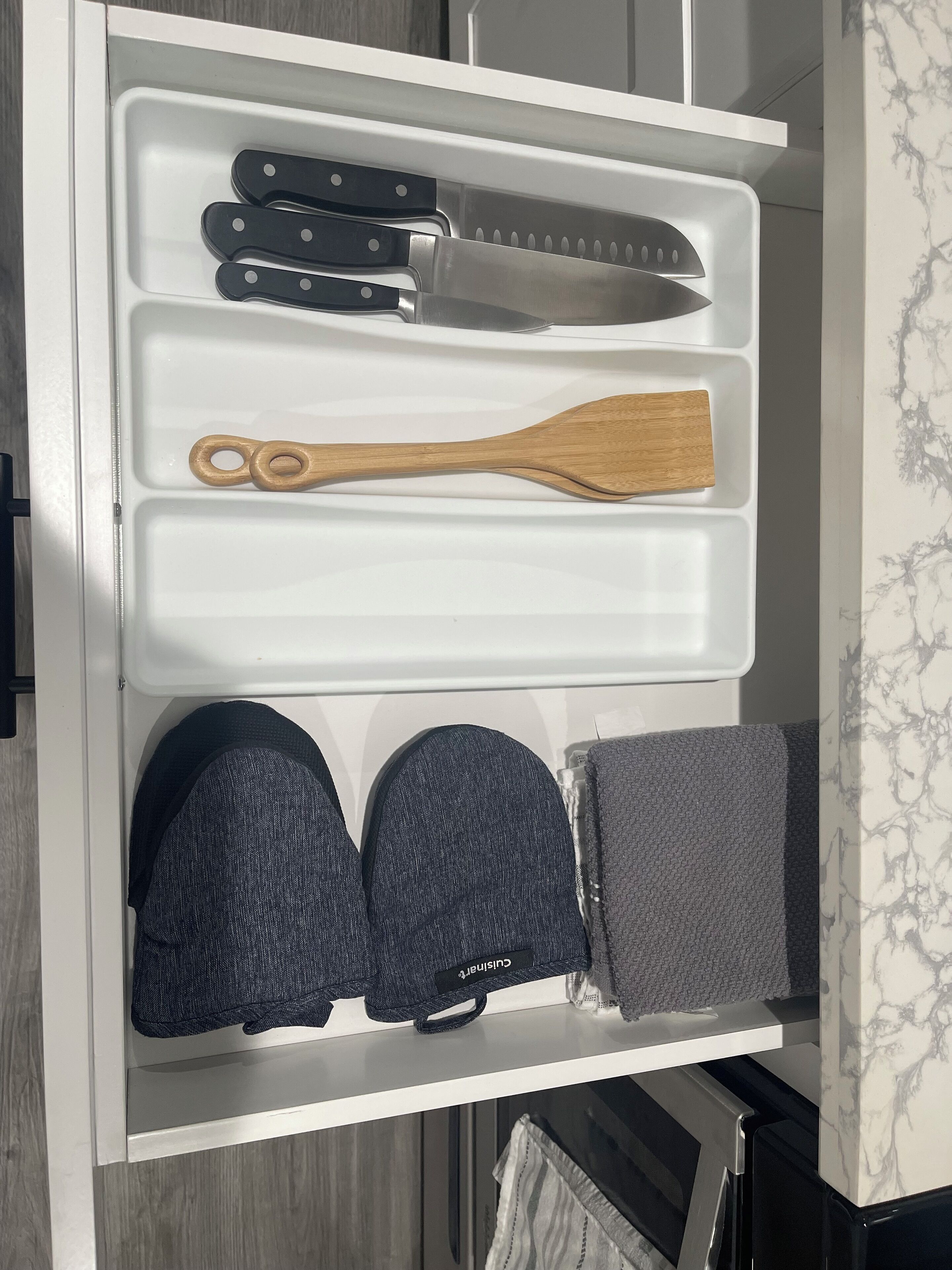 A kitchen drawer with knives, a wooden spatula, and two pairs of oven mitts.