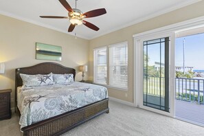 A bedroom with a bed, ceiling fan, artwork, and a view through the window.