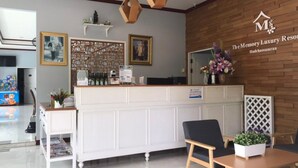 Un espace d’accueil avec un comptoir de réception blanc, une chaise noire et une table en bois sur laquelle est posée une plante en pot. Des tableaux encadrés ornent le mur, et une enseigne est placée au-dessus du comptoir.