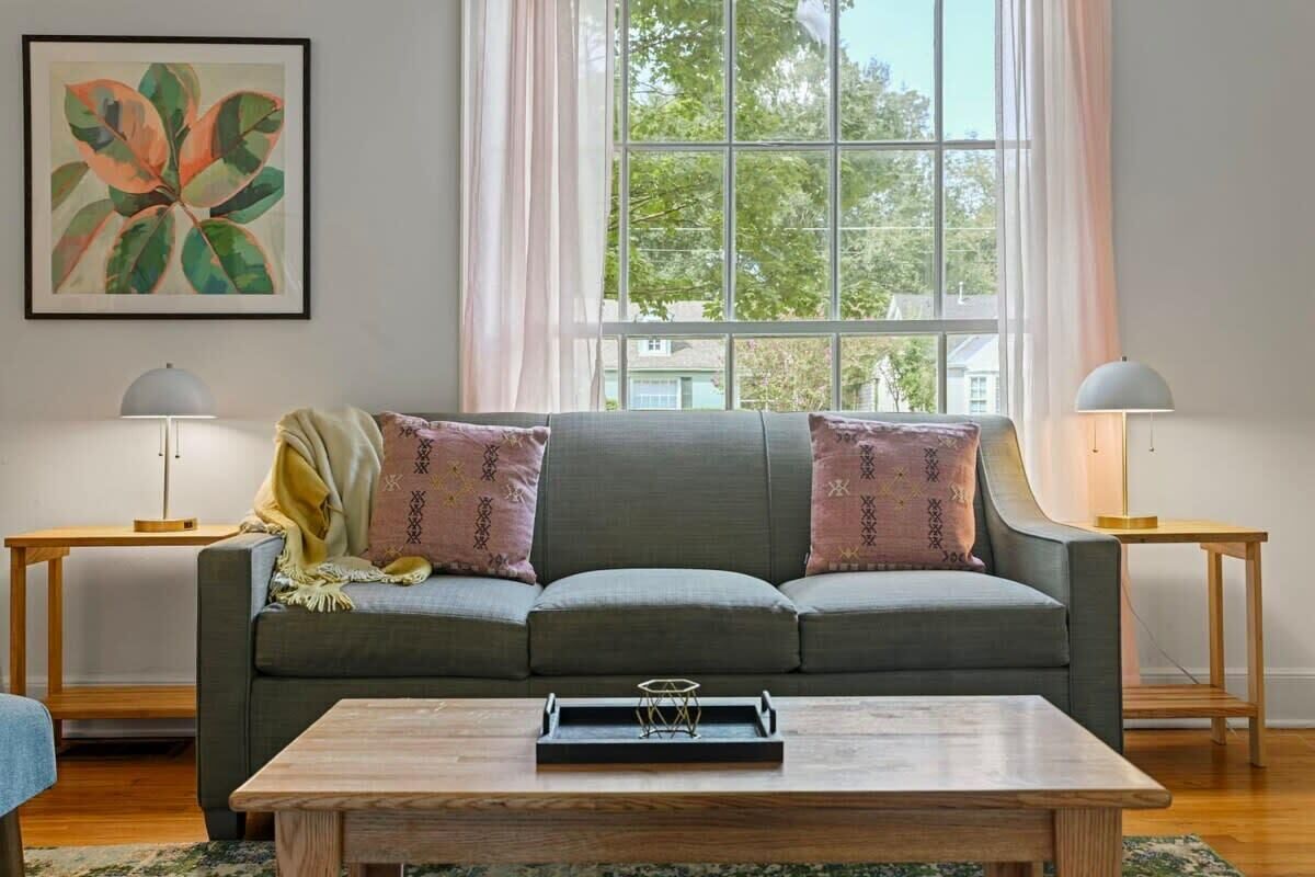 Ein Wohnzimmer mit grauem Sofa, zwei Beistelltischen mit Lampen, einem Holztisch als Couchtisch und einem großen Fenster mit Vorhängen.