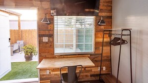 Ein kleines Zimmer mit Holzwand, einem Fenster mit Jalousien, einem kleinen Tisch, einem Stuhl und einem über dem Fenster angebrachten Fernseher.