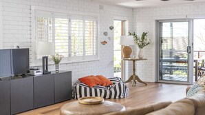 A modern living room with a white brick wall, a flat-screen TV, a striped bean bag, a leather ottoman, and a sliding glass door leading to an outdoor area. - Bimbajong Studio - Pet Friendly- Oceanstays (Yamba)