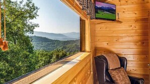 A wooden cabin with a television mounted on the wall, a leather chair with a cushion, and a view of a forested landscape. - Private Mtn Views|Movie Theater|Hot Tub|Pool Table (Sevierville)