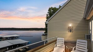 En balkon med to hvide stole, der kigger ud over en rolig sø og et havneområde i solnedgang.