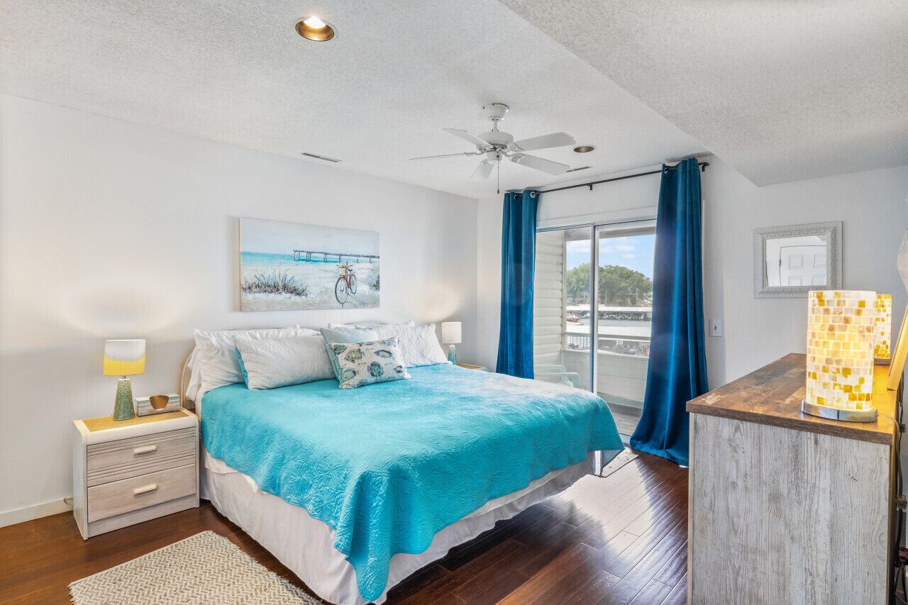 Un dormitorio con cama, mesita de noche, ventilador de techo, ventana con cortinas y vistas al exterior.