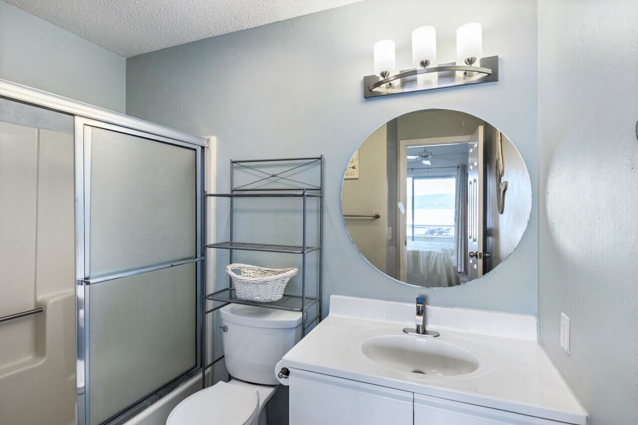 A bathroom with a glass shower enclosure, a toilet, a sink with a mirror above, and a wall-mounted shelf with a basket.