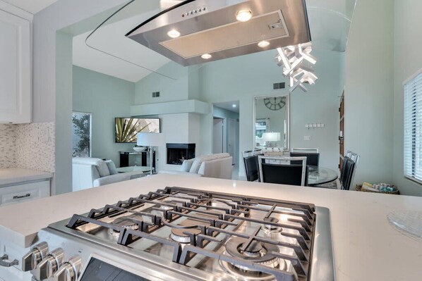 A modern kitchen with a central island, stainless steel appliances, and a dining area with a round table and chairs. - Desert Refresh | 3BR Santa Rosa Cove (La Quinta)