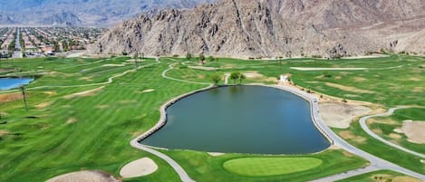 A golf course with a pond, surrounded by mountains and residential areas.