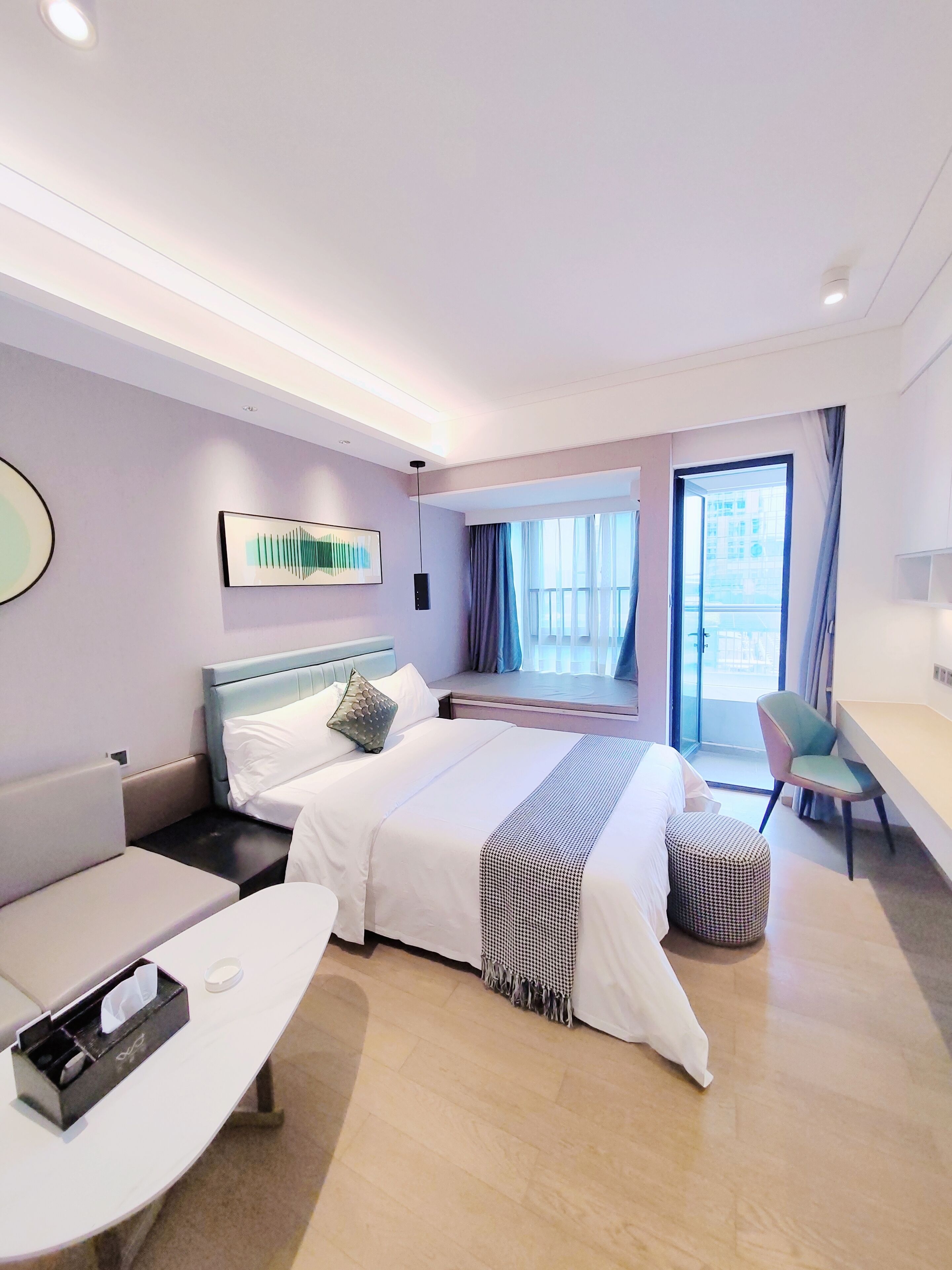 A modern hotel room with a large bed, a desk, and a chair. There is a window with curtains, a wall-mounted artwork, and a ceiling light.