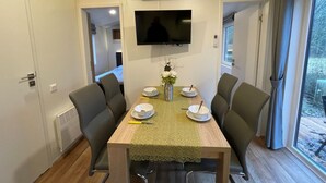 Un comedor con una mesa dispuesta para cuatro personas, rodeada de sillas, un televisor de pantalla plana en la pared y un ventanal con cortinas.