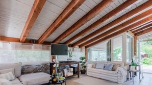 A spacious living area with a high ceiling and exposed wooden beams, a large flat-screen TV, a sofa, a coffee table with a tray of drinks, and a view of greenery outside. - Vacation home Serena by Interhome (Rio Marina)