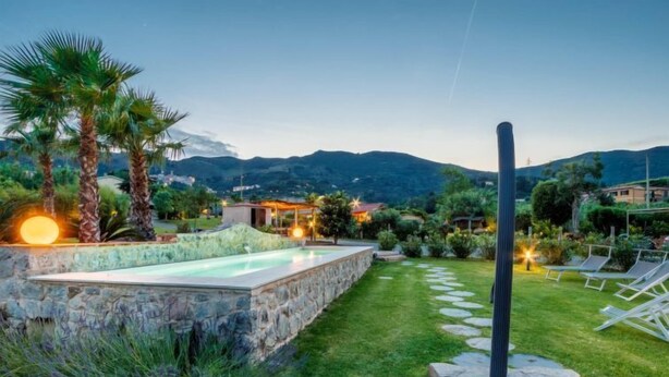 A landscaped garden with a swimming pool, stone walls, and a pathway leading to a seating area with lounge chairs. - Vacation home Serena by Interhome (Rio Marina)
