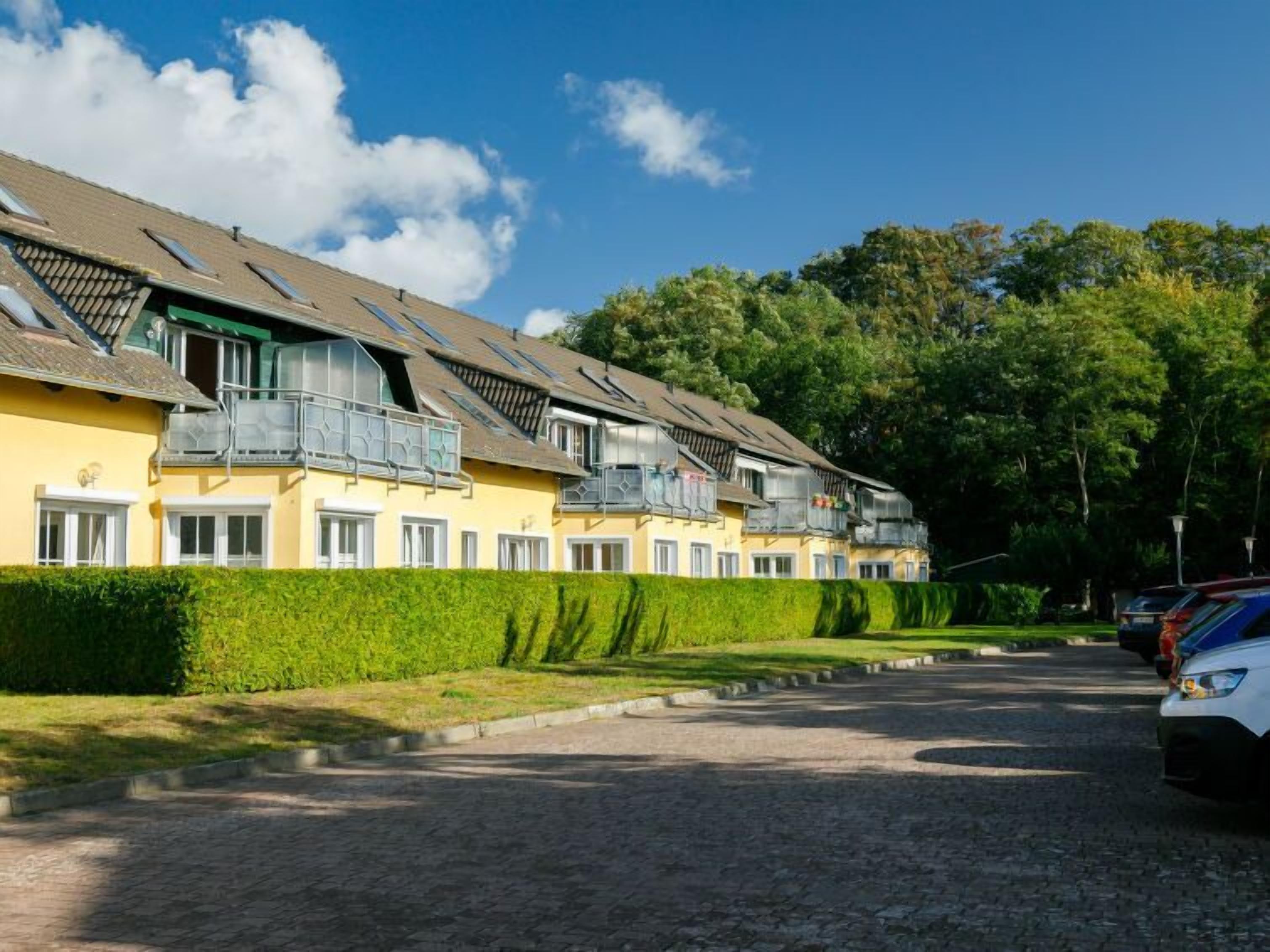 A row of yellow buildings with balconies, a paved driveway, and a hedge.