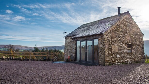 A stone cottage with a sloped roof, large glass doors, and a gravel driveway. - The Hidden Place (Penrith)