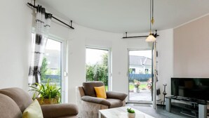 A modern living room with a sofa, armchair, and coffee table, a flat-screen TV, and large windows offering a view of the outdoors. - Schneider by Interhome (Zinnowitz)