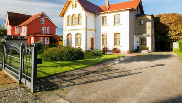 Een groot, tweelaags huis met een rood dak en witte muren, omgeven door een verzorgd gazon en een metalen hek.