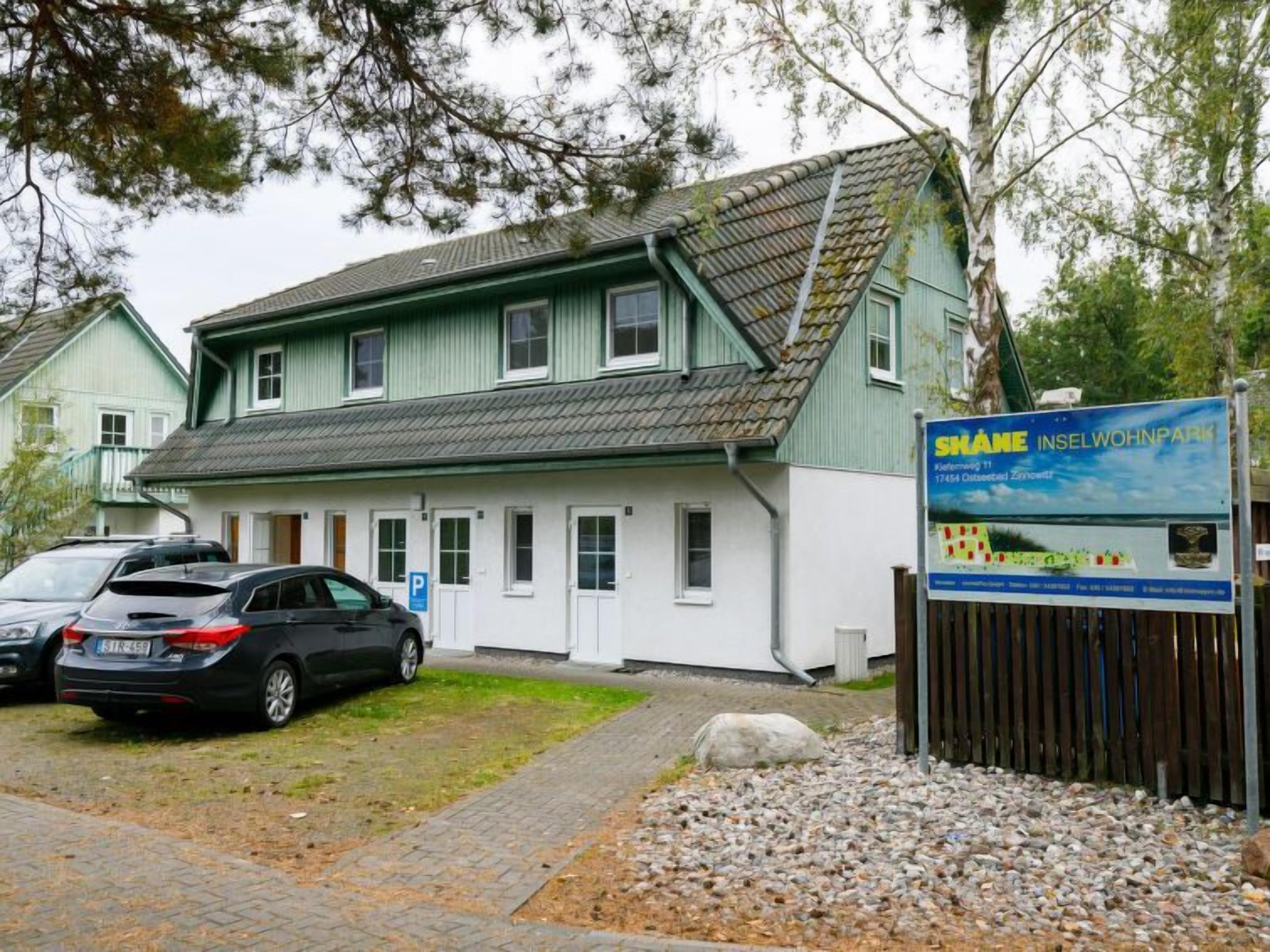 En toetagers bygning med en grøn facade og et skråt tag. Der er et skilt med tekst og et logo, samt en parkeringsplads med biler.