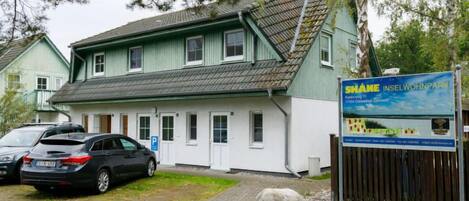En toetagers bygning med en grøn facade og et skråt tag. Der er et skilt med tekst og et logo, samt en parkeringsplads med biler.