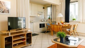 A modern living room with a flat-screen TV, wooden entertainment unit, dining table with chairs, and a kitchen area with white cabinets and appliances. - Zur Düne II by Interhome (Zinnowitz)