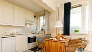 A modern kitchen with white cabinets, a dining area with wicker chairs, a wooden table, and a window with curtains. - Zur Düne II by Interhome (Zinnowitz)